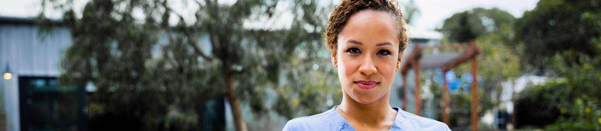 Woman in scrubs looks at the camera, standing in front of trees and greenery.