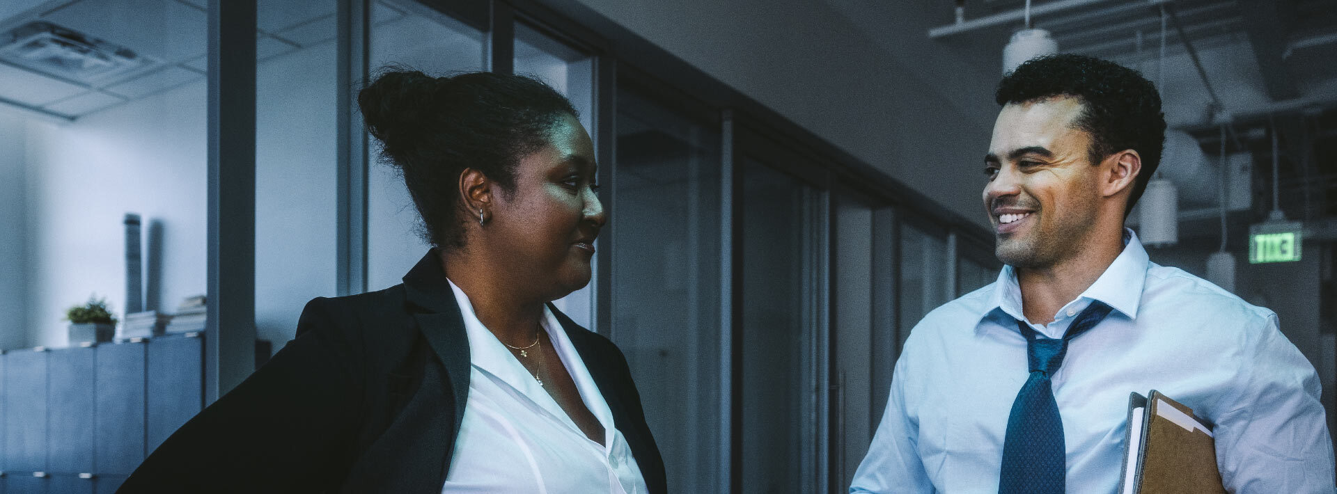 A man and woman, both professionally dressed, talk in an office