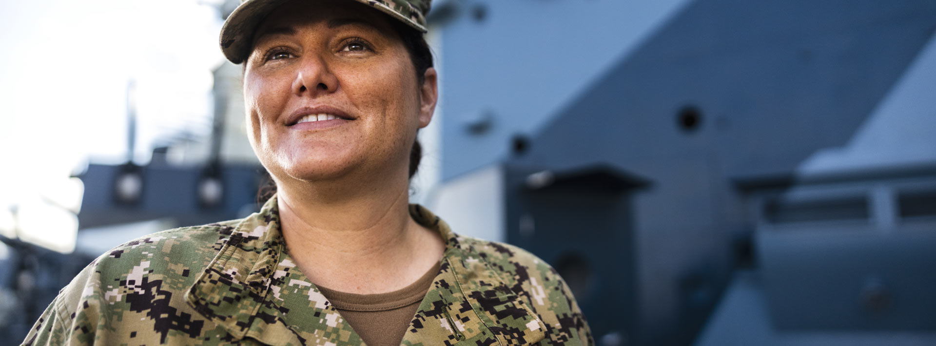 A woman in military uniform stands in front of buildings and looks off into the distance.