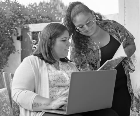 Two women study a notebook while one types on a laptop